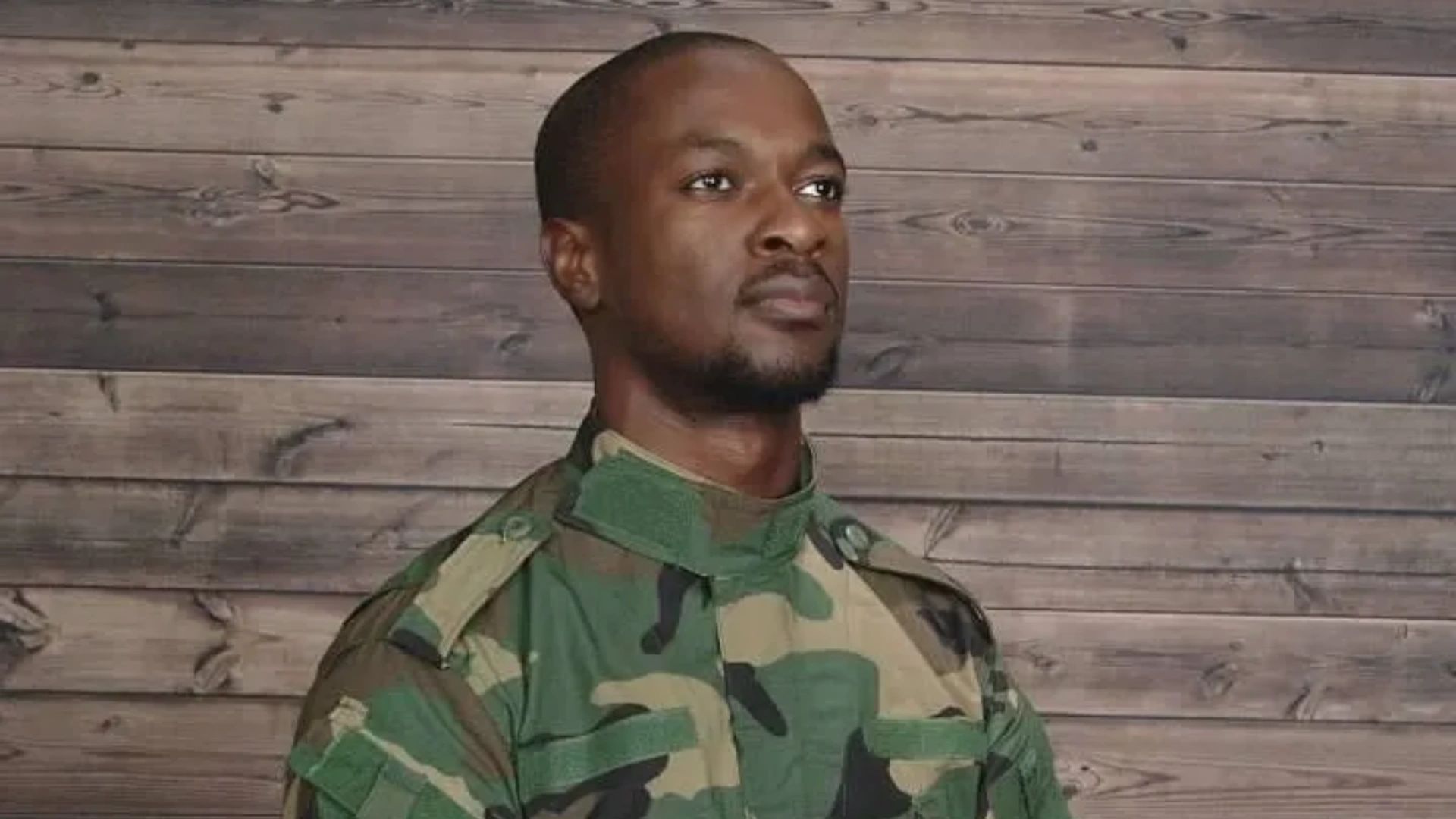 Man in camouflage military uniform standing against wooden wall.