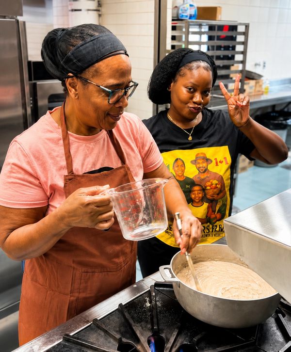 Two women cooking together in a kitchen, one stirring a pot and the other making a peace sign.
