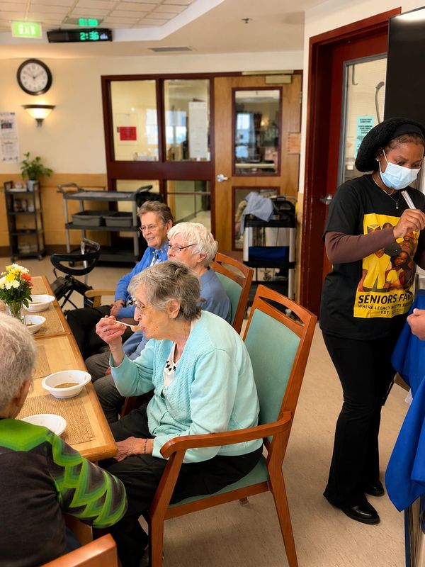 Elderly people eating together in a communal dining area with a caregiver nearby.