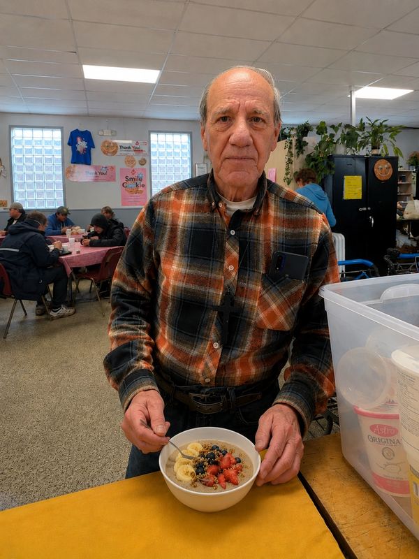 Elderly man holding a bowl of oatmeal with fruit in a community dining area.