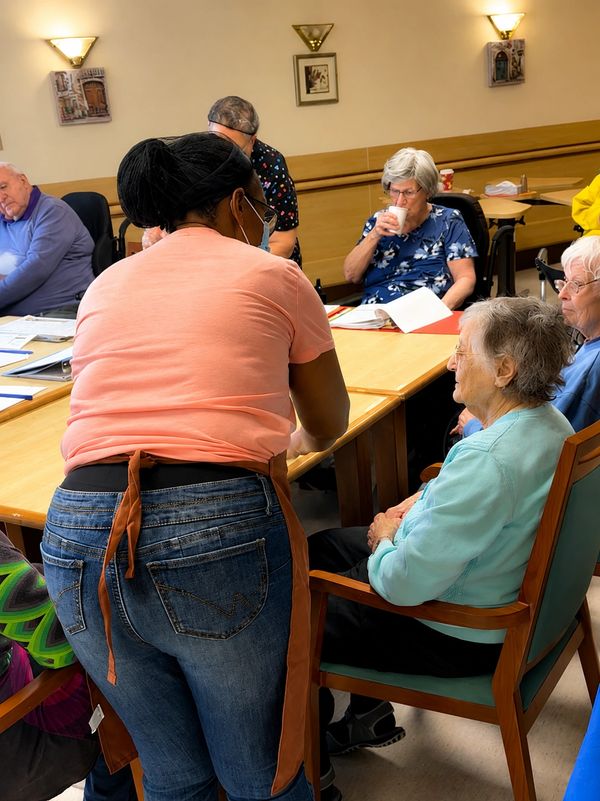 Caregiver assisting elderly woman in a communal room with others.