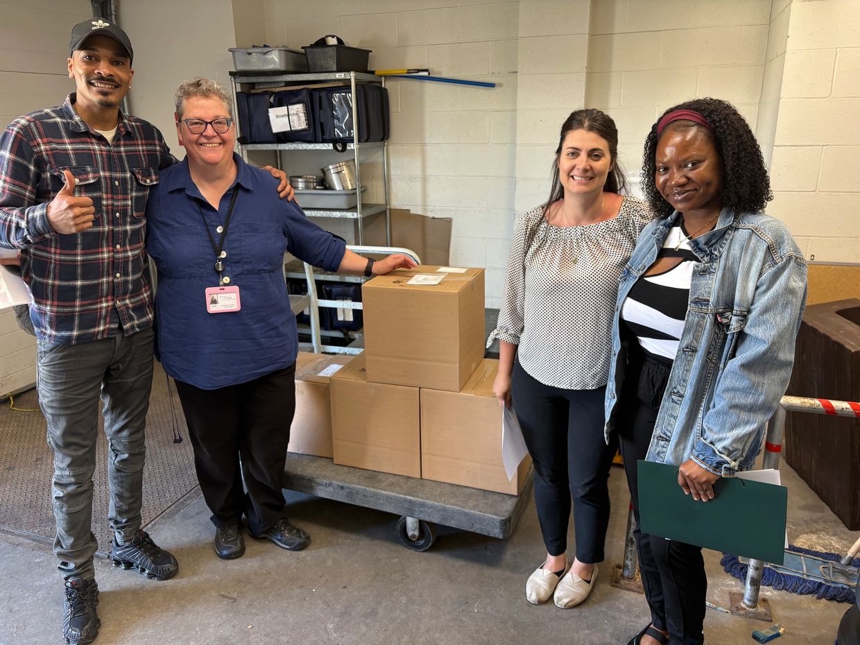 Four people smiling by a cart with stacked cardboard boxes in a storage room.