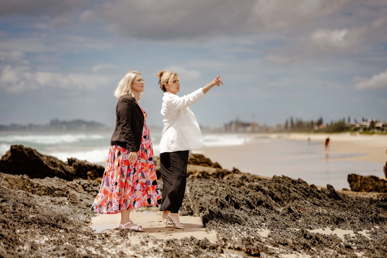 Two women stand on rocky beach, one pointing towards the horizon under a cloudy sky.