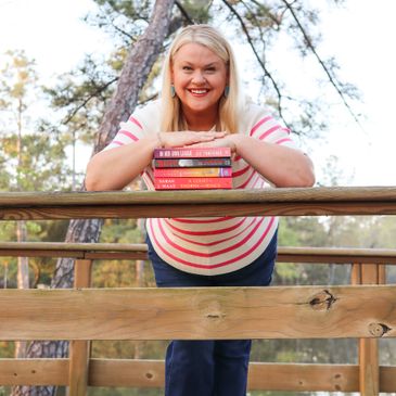 Smiling woman leaning on a wooden railing with a stack of books outdoors.