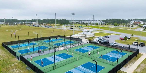 Aerial view of multiple blue pickleball courts with players and surrounding parking.
