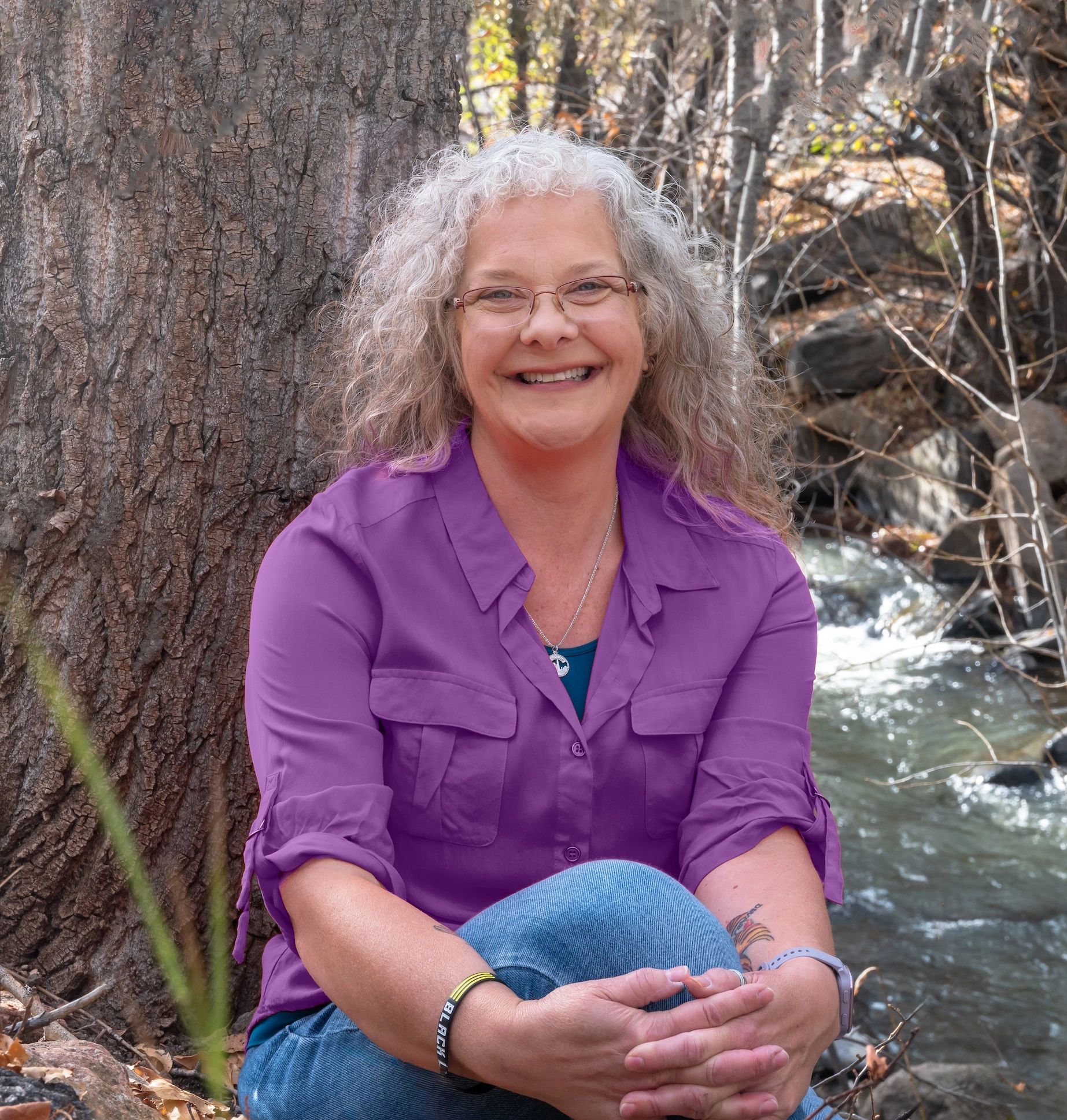 Smiling woman with curly gray hair in a purple shirt sitting outdoors by a tree and stream.