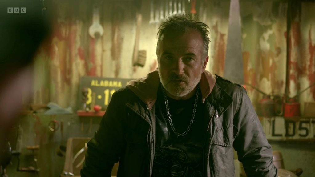 A man with a chain necklace in a rustic, dimly lit room.