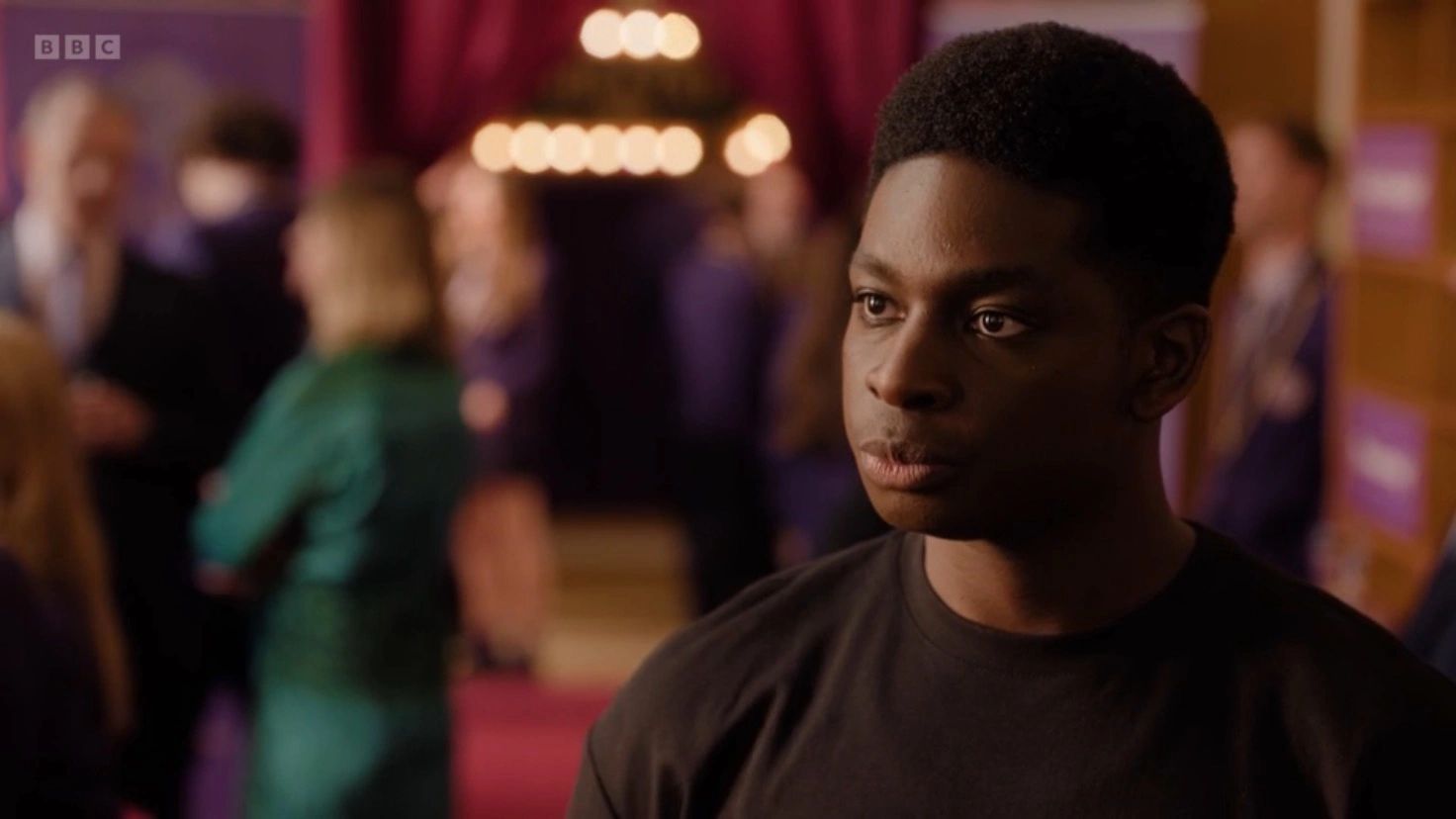 Serious young man in black shirt amidst a blurred crowd at a BBC event.