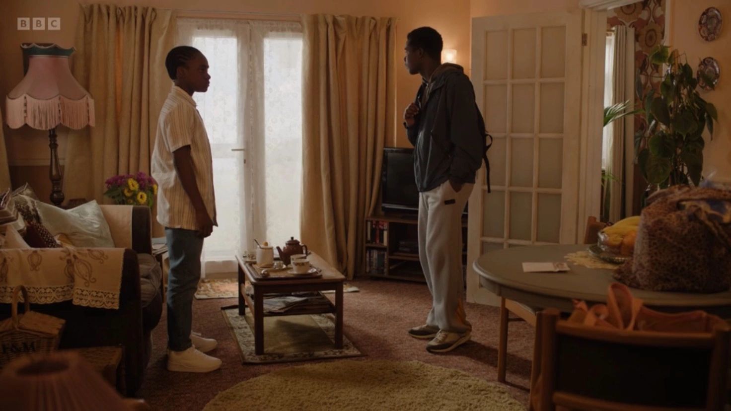 Two young men stand and talk in a warmly lit living room.