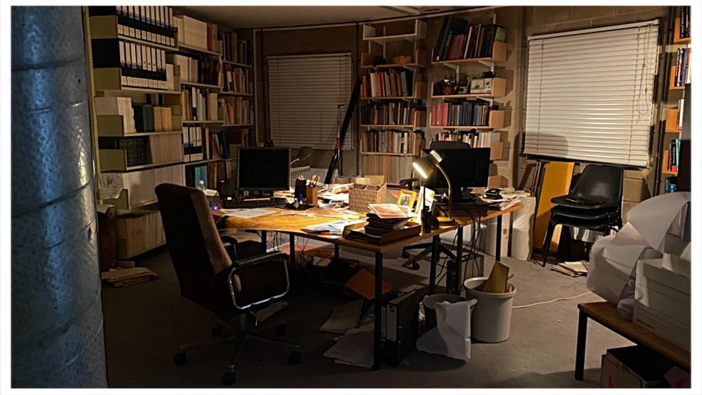 Dimly lit cluttered study room with books and computer screens.