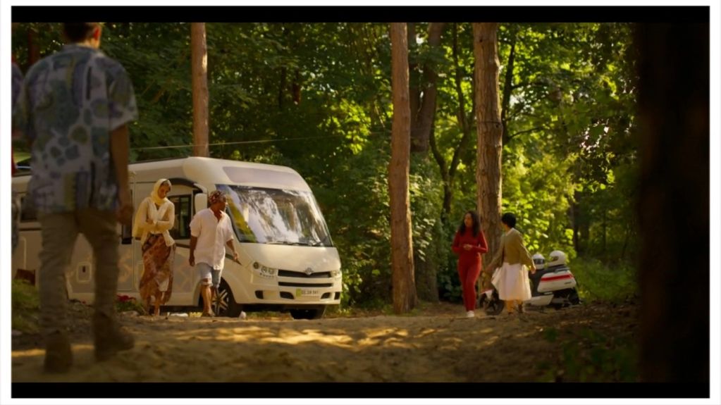 People walking near a white camper in a forest.