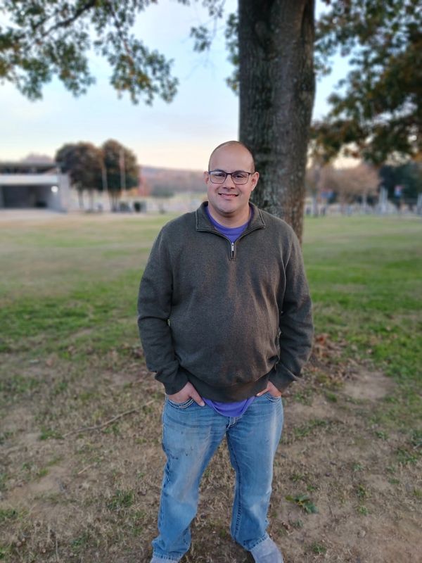 Man wearing glasses and a sweater stands outdoors near a tree.