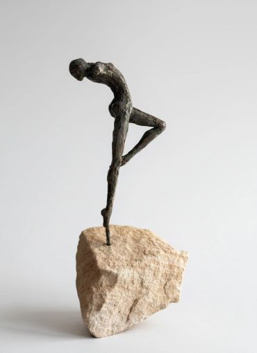 Abstract bronze sculpture of a slender human figure balancing on one leg atop a rock.