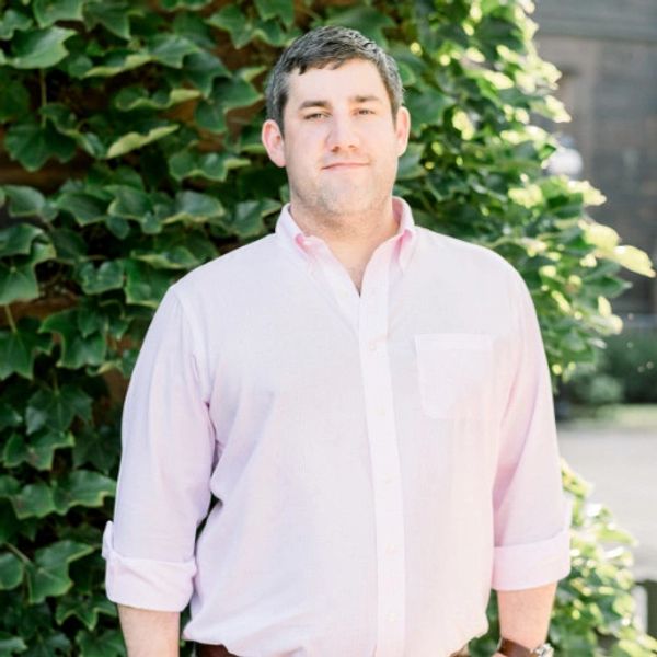 Man in light pink shirt standing outdoors against leafy background.