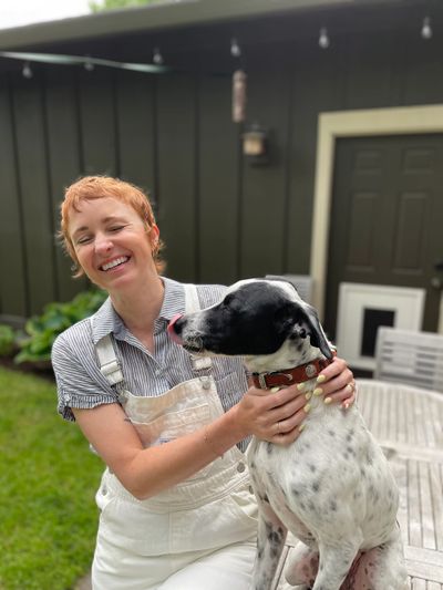 Smiling person in overalls cuddling a black and white dog outdoors.