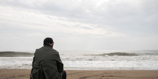 An older man sitting on a weathered post at the edge of the ocean, as the waves roll in.