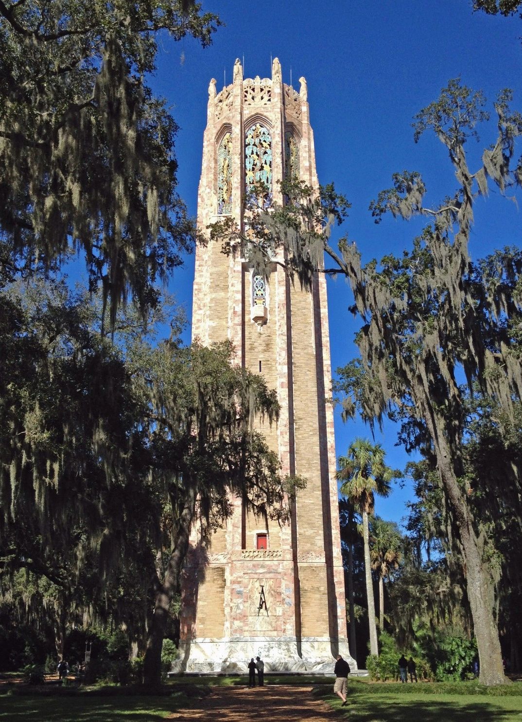 A tall, tan-colored tower with stained glass windows surrounded by trees and grass