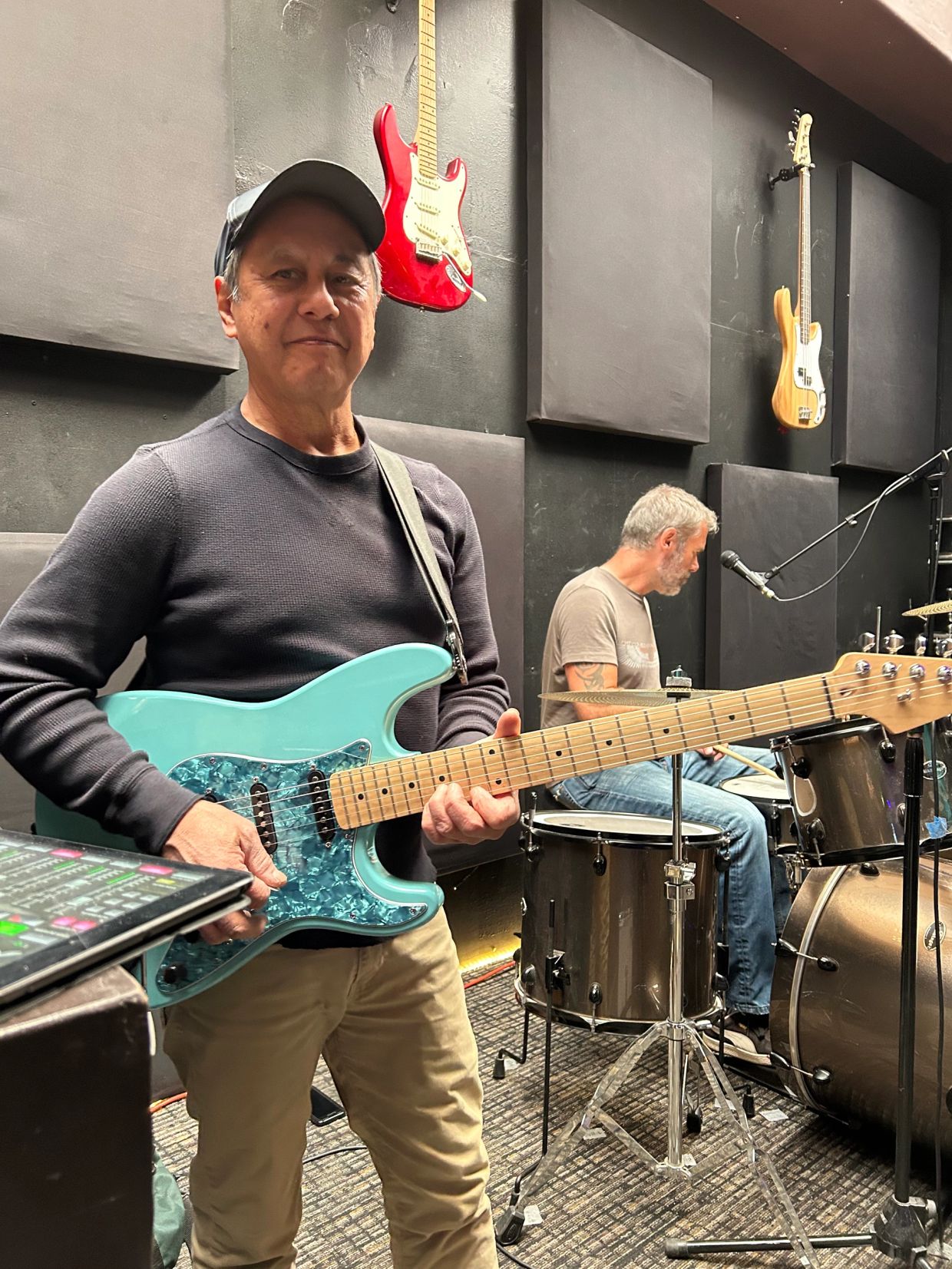 Man with teal electric guitar, drummer in background.