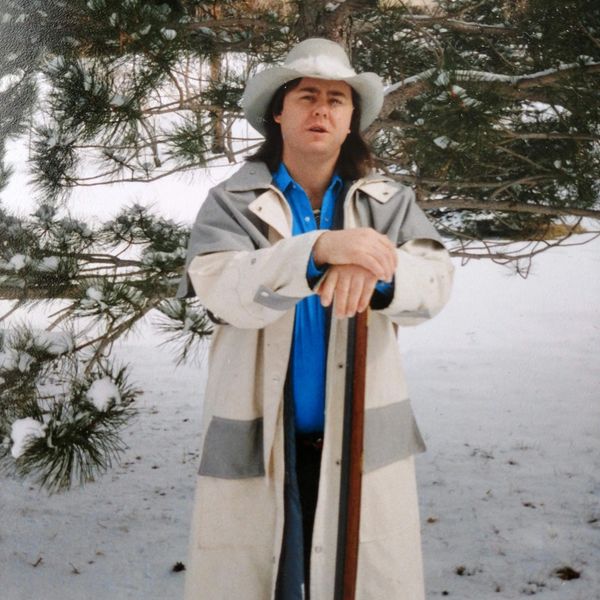 Man in a white coat and hat standing in snowy woods, holding a walking stick.