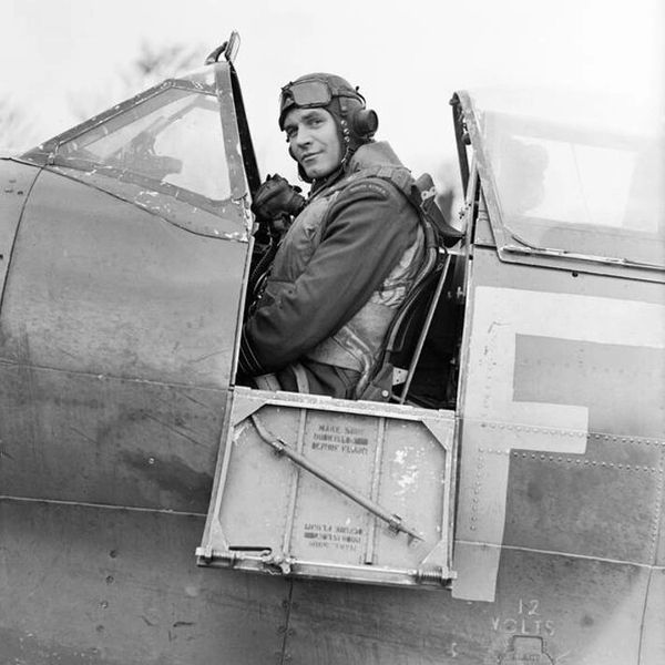 A pilot in vintage flight gear sits in the cockpit of a WWII-era fighter plane.