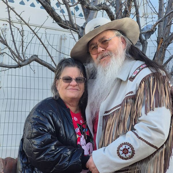 A couple embraces outdoors, one wearing a fringed jacket and hat.