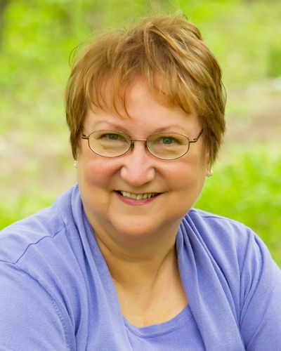 Author Joyce Proell wearing a lavender sweater set, she has short layered hair and glasses.