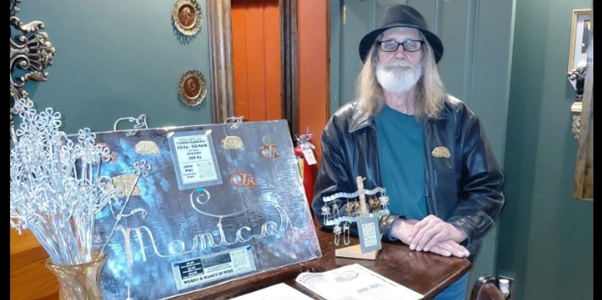 Man with long hair and beard standing by a table with handmade metal art and jewelry.
