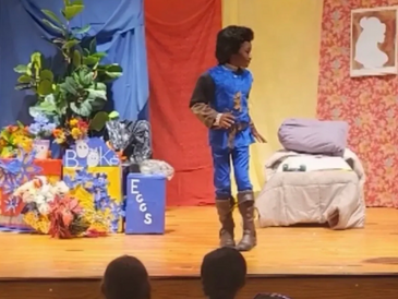 A child in a blue costume performs on stage in a colorful play setting.