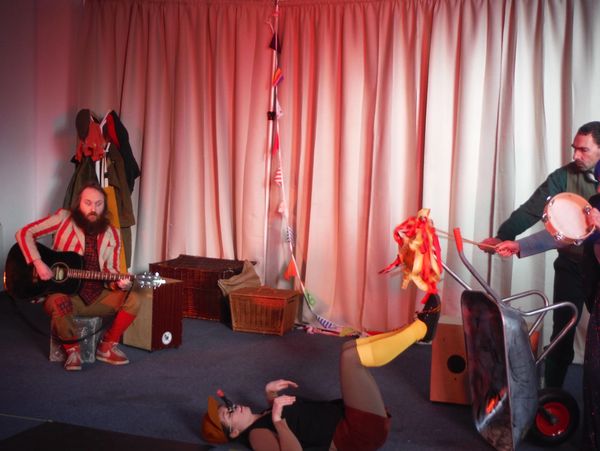 Three performers engaging in a quirky, colorful indoor act with instruments and props.