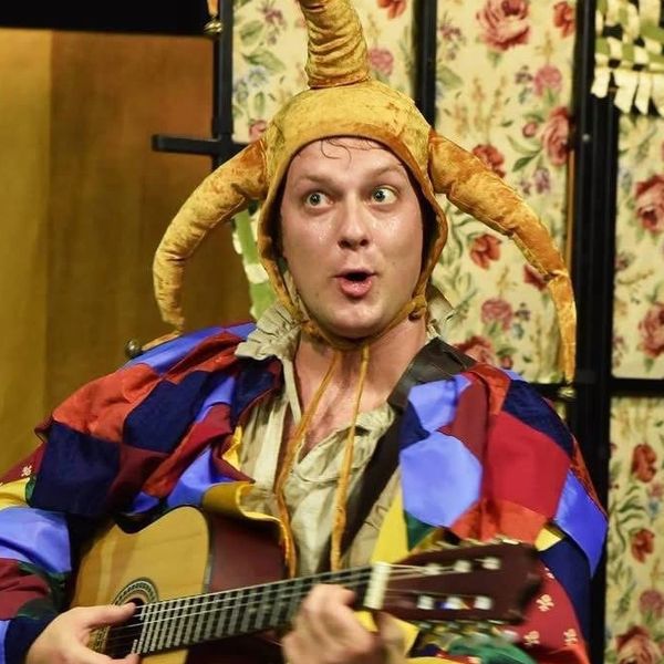 A man in a colorful jester costume playing guitar with an expressive face.