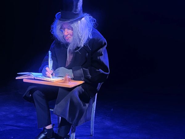 Person in a tall hat and dark coat sitting at a small desk writing.
