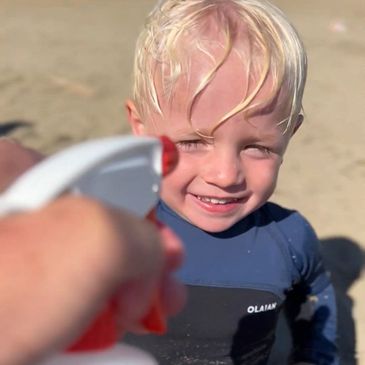 A young boy in a wetsuit smiles as water is sprayed towards him at the beach.
