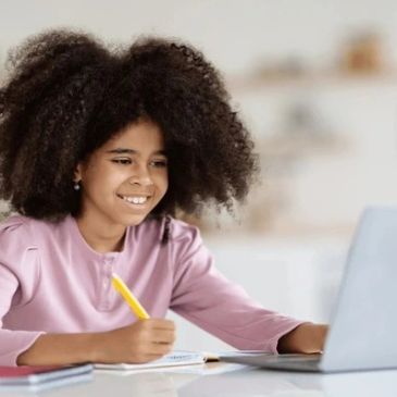 Smiling girl with curly hair using a laptop and writing in a notebook.