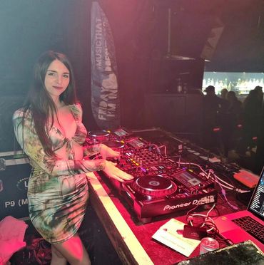 A female DJ stands behind a Pioneer DJ console at a club, smiling.