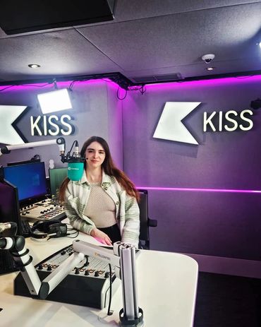A young woman in a radio studio with "KISS" signs and professional equipment.