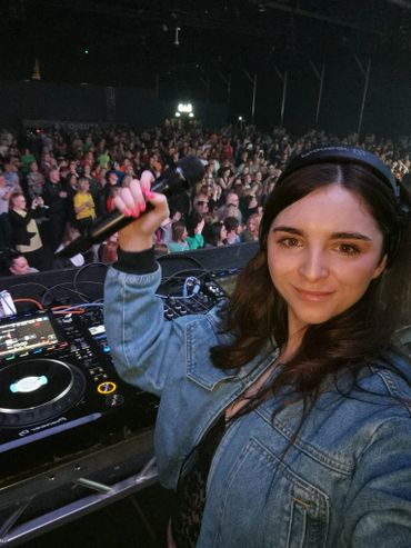 Female DJ holding a microphone, smiling at a crowded music event.
