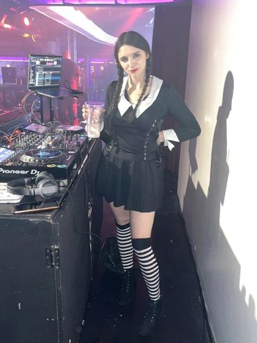 Woman in black and white outfit with striped socks stands by DJ equipment holding a drink.