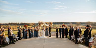 Outdoor wedding ceremony with bride, groom, and guests on a sunny day.