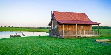 Wooden cabin with a red roof beside a pond in a vast green field.