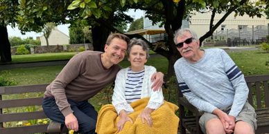Three people sitting closely on a bench outdoors, smiling warmly.