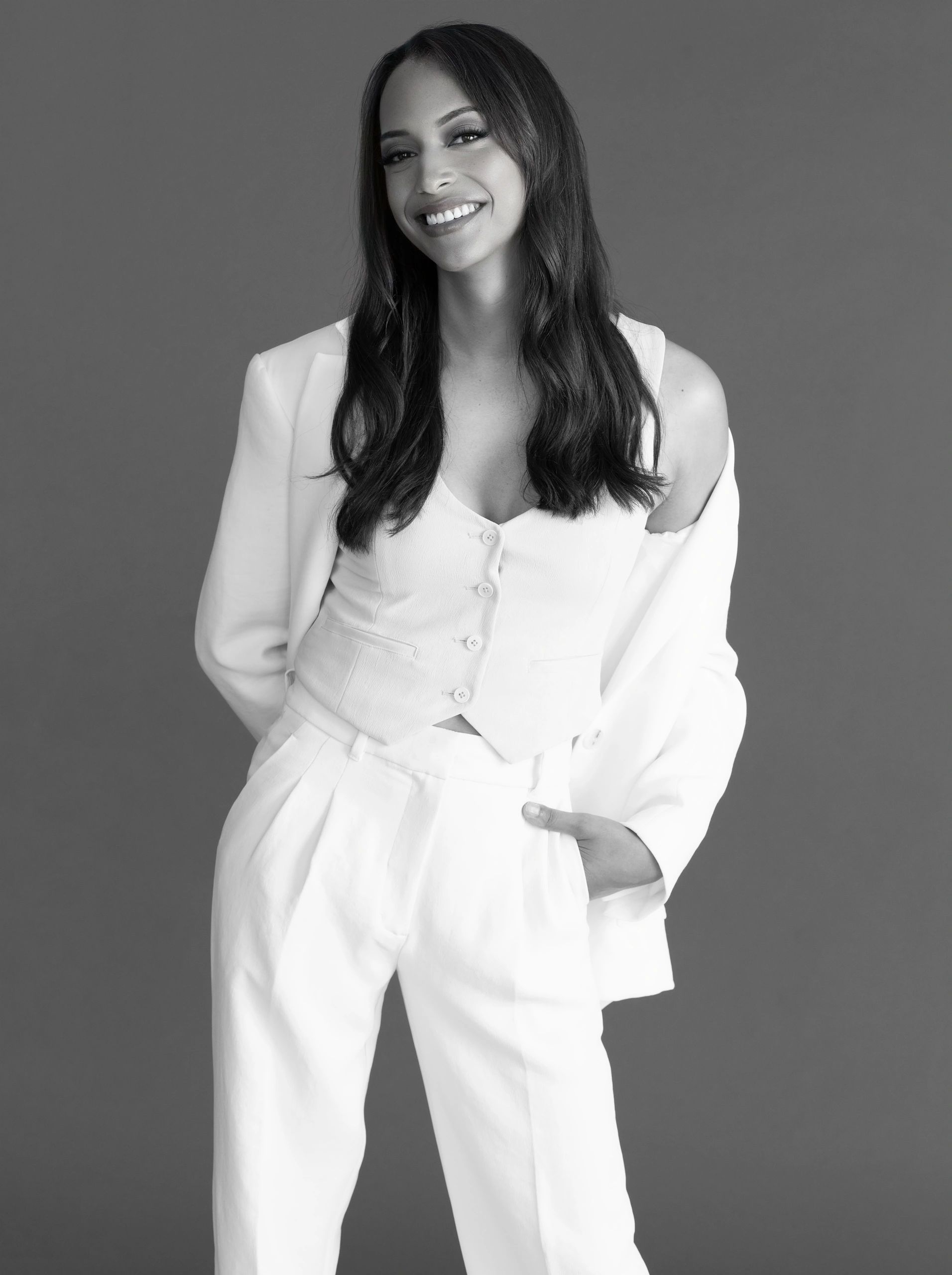 Confident woman in stylish white outfit smiling against gray background.