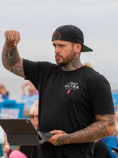 Man with tattoos and cap holding a laptop, flexing his arm outdoors.