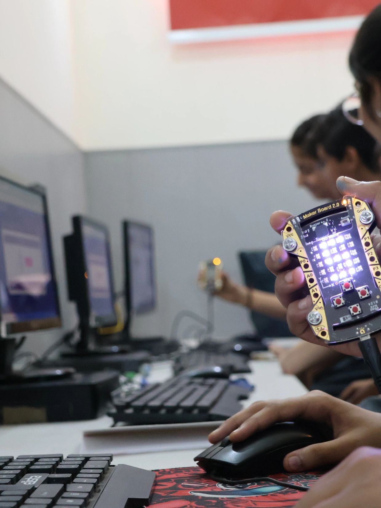Person holding a Maker Board 2.0 near a computer mouse in a tech workspace.