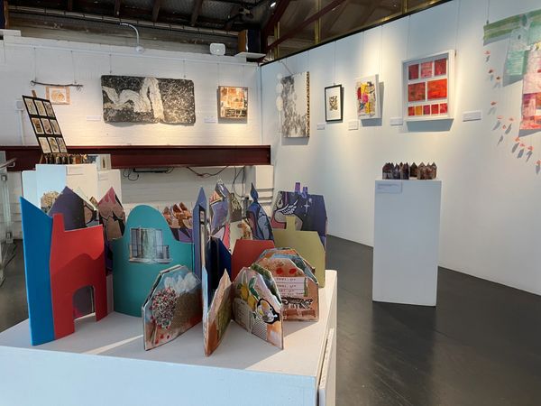 Art gallery displaying colorful 3D paper houses and framed artwork on white walls.