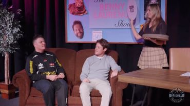 Two men seated on a couch while a woman stands holding a tray.