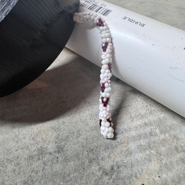 A beaded bracelet with white and purple beads on a concrete surface near a white pipe.
