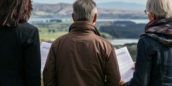 Three people stand outdoors holding papers, overlooking a scenic landscape with hills and water.