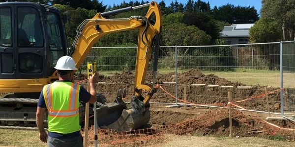 Construction worker using a surveying tool near an excavator at a digging site.