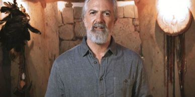 Man with gray beard in a rustic room with stone wall and warm lighting.