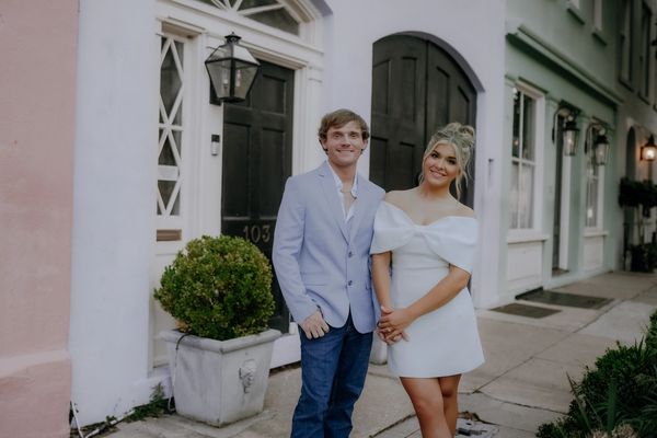 A couple dressed elegantly stands smiling on a charming street with pastel-colored buildings.